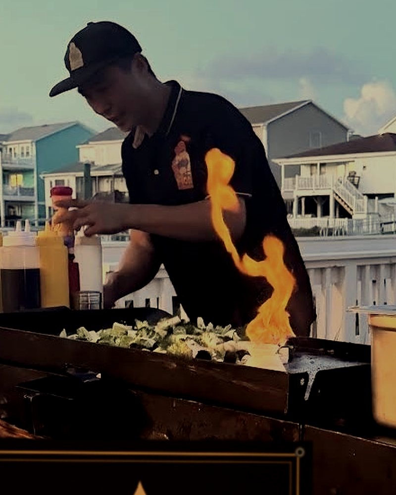 Private hibachi chef flame-cooking vegetables on a waterfront deck, beach houses behind